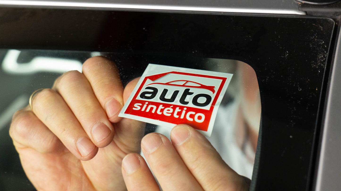 Manos colocando una pegatina de cambio de aceite Autosintetico® en el parabrisas, un adhesivo de mantenimiento duradero y resistente, similar a las pegatinas autoestáticas utilizadas en talleres.